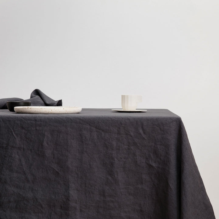 The Linen Tablecloth in Slate styled with a white ceramic plate and white cup and saucer. Available in Small 150cm x 240cm & Medium 180cm x 300cm.