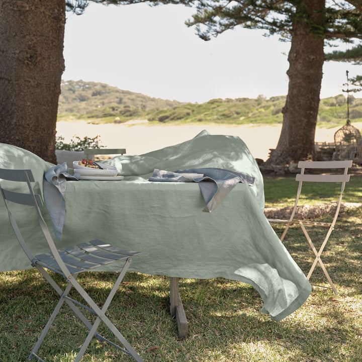 A rectangular table in an outdoor setting is dressed with a Linen Tablecloth in Sage and Linen Napkins in Bluestone. Available in Small 150cm x 240cm & Medium 180cm x 300cm.