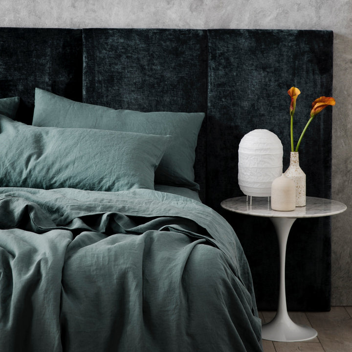 A bed dressed in Bluestone bed linen, styled with a velvet headboard, modern bedside table, small ceramic vases and small white lantern. Available in Double, King, Super King.