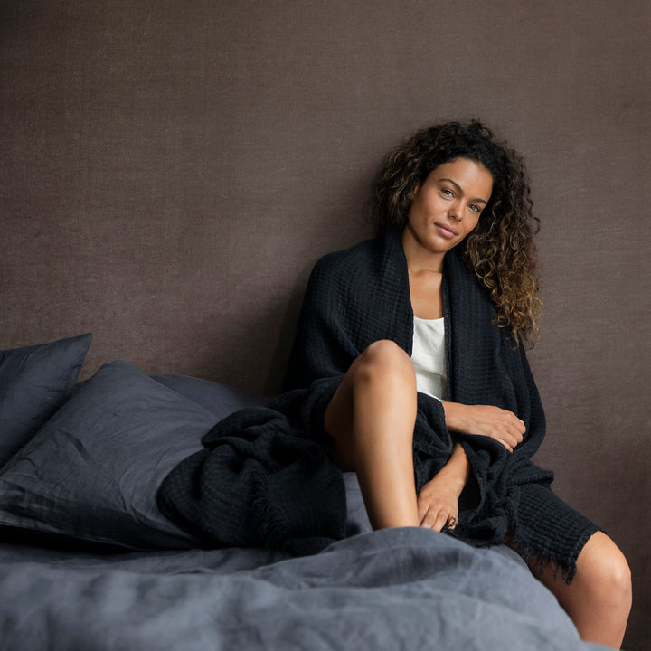 A woman is sitting on a bed dressed in Slate bedlinen, she is wearing a Black Linen Waffle Robe. Available in Standard & King.