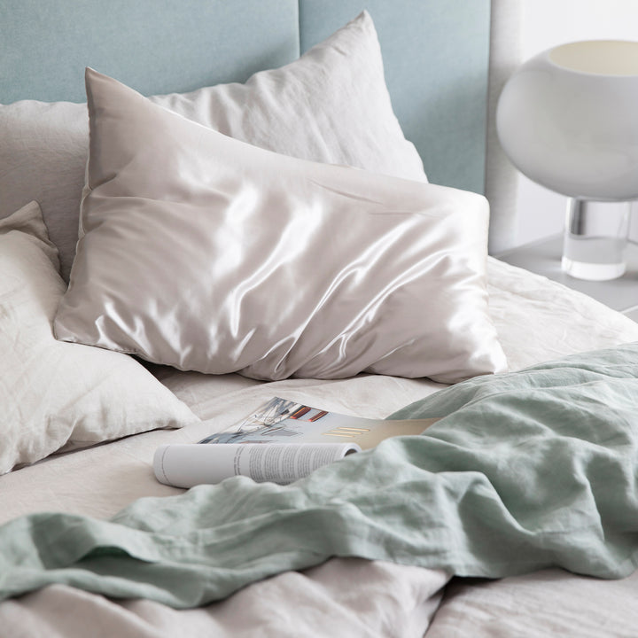 Bed styled with the Smoke Gray Duvet Cover set, a Smoke Gray Fitted sheet, Sage Flat Sheet with Border and a Smoke Gray Silk Linen Flip Pillowcase. Also featured is an open magazine positioned atop the sheets.