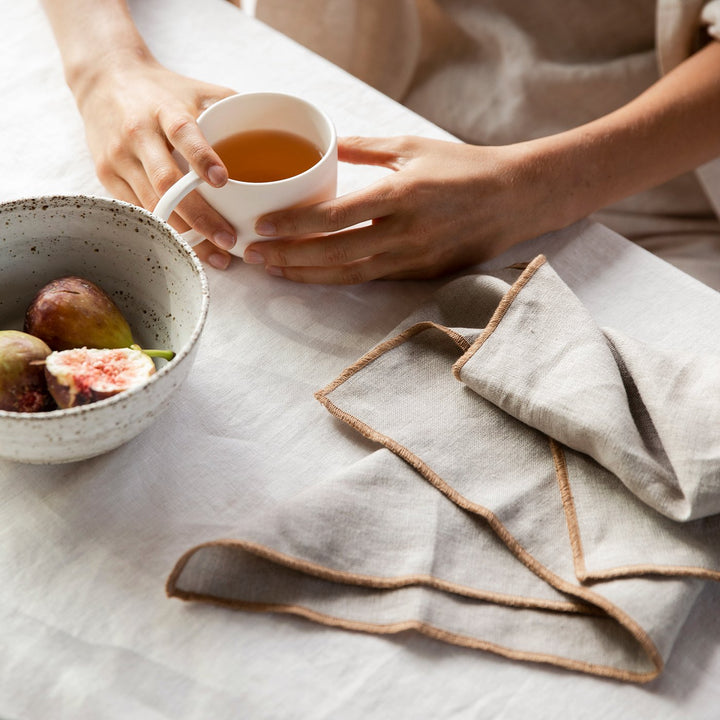 Model is sitting at the table with both hands cupping a mug of tea. There is a speckly ceramic bowl of three figs in front of her hands and a Cara Edged Napkin in Natural placed to her left.