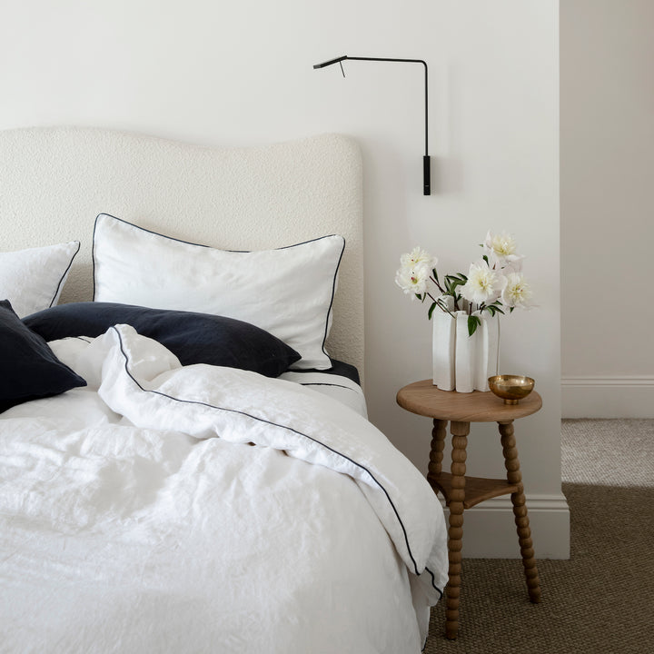 A cream bed dressed in White and Navy Piped bedlinen with a Navy Fitted Sheet and Pillowcases is styled with a side table and vase of flowers. Available in Standard & King.