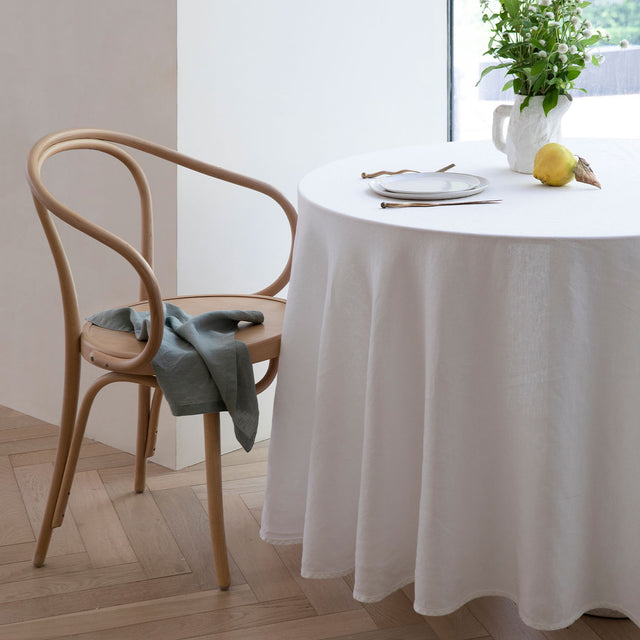 A table dressed in a White Linen Tablecloth, styled with a wooden chair and napkin. Available in Small 150cm x 240cm & Medium 180cm x 300cm.