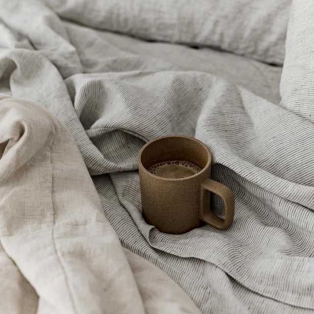 A close up of a bed dressed in Pinstripe and Natural bed linen, styled with a brown coffee mug