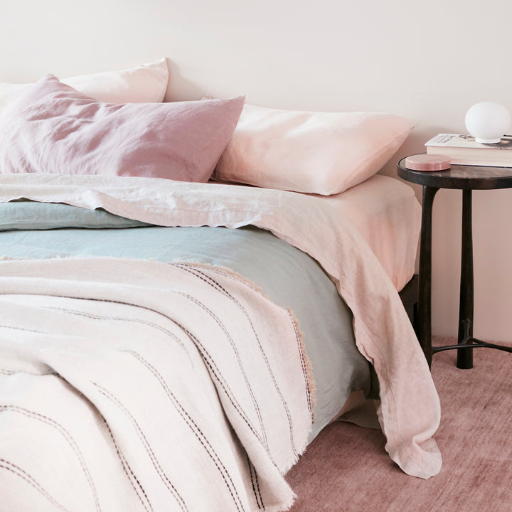 A bed dressed in a Sheet Set in Blush with a Pillowcase in Dusk, a Duvet Cover in Sage and a Mira Ana Bedcover. A black side table holds a small lamp and magazines. Available in Standard & King.