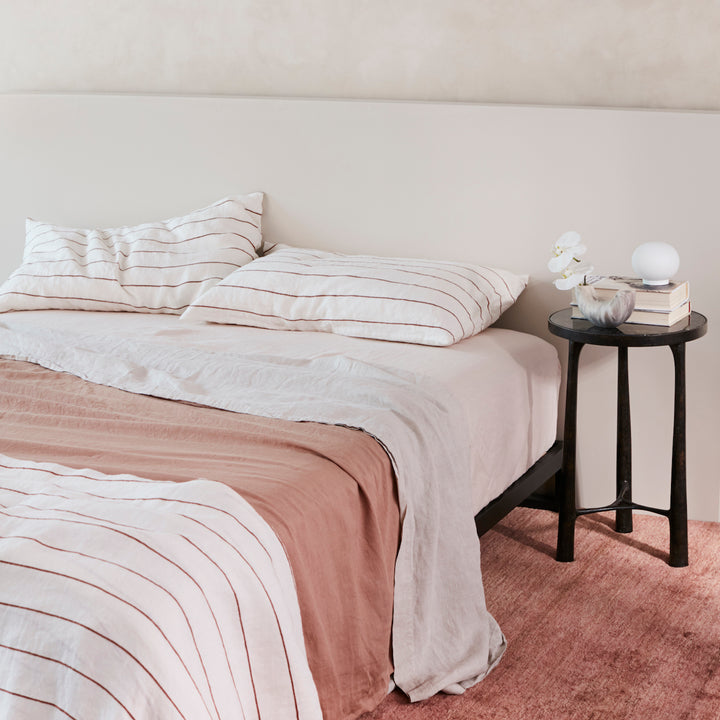 A bed dressed Blush, Cedar Stripe, Fawn and Smoke Grey bed linen, styled with a black bedside table, a pile of books and a small vase with flowers. Available in Double, King, Super King.