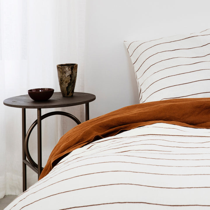  A close up of a bed dressed in Cedar Stripe and Cedar bed linen, styled with a modern bedside table, ceramic bowl and vase