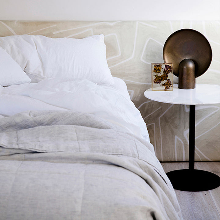 A bed dressed in White bedlinen, with a Pinstripe Quilt Cover, is styled against a printed bedhead and a side table with brass objects. Available in Single, Double, King, Super King.