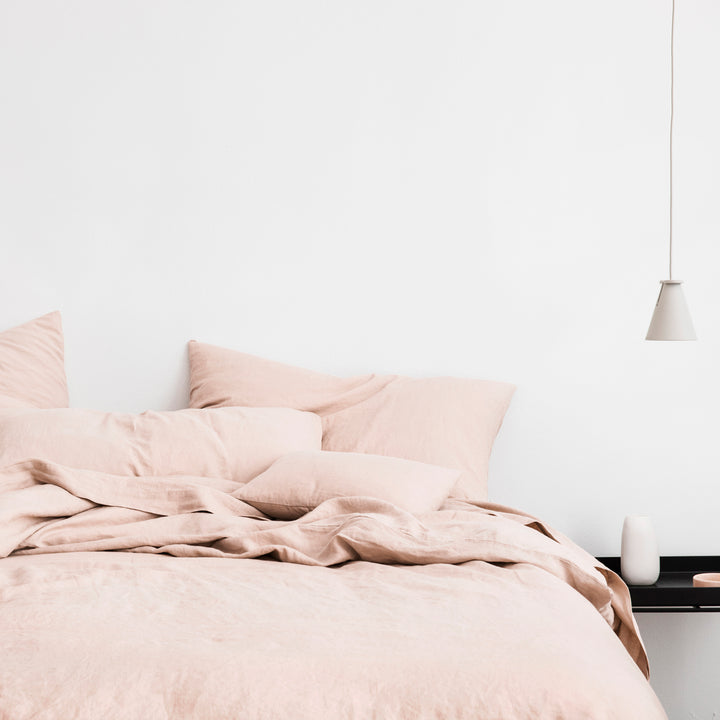 A bed dressed in Blush bedlinen styled with a black side table holding a white vessel. A simple white pendant lamp hangs above the table. Available in Standard & King.