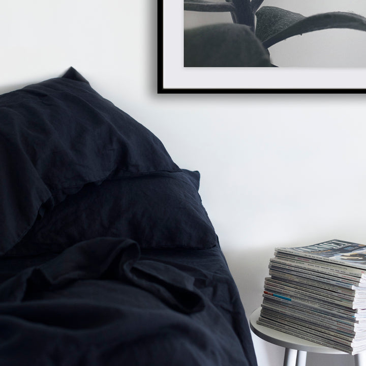 A cropped photo of a bed dressed in Navy bed linen. There is a white bedside table next to the bed with a stack of magazines on top of it. Available in Double, King, Super King.