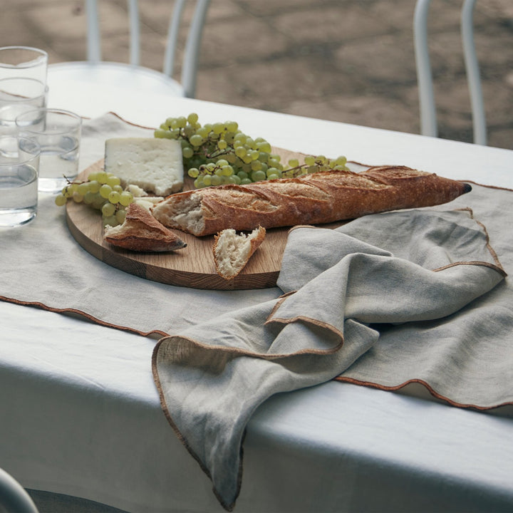 An outdoor table dressed in the Cara Panel Tablecloth, Cara Edged Table Runner in Cedar and a Cara Edged Napkin in Natural. On top of the table linen is a round wooden board of cheese, grapes and a baguette.