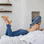 A female model is lying on a bed styled with a Linen Duvet Cover Set in Pinstripe and a Linen Flat Sheet with Border and Linen Fitted Sheet in White. The model is reading a book and has a Denim Bath Towel wrapped around her hair, and is also wearing a pair of denim jeans.