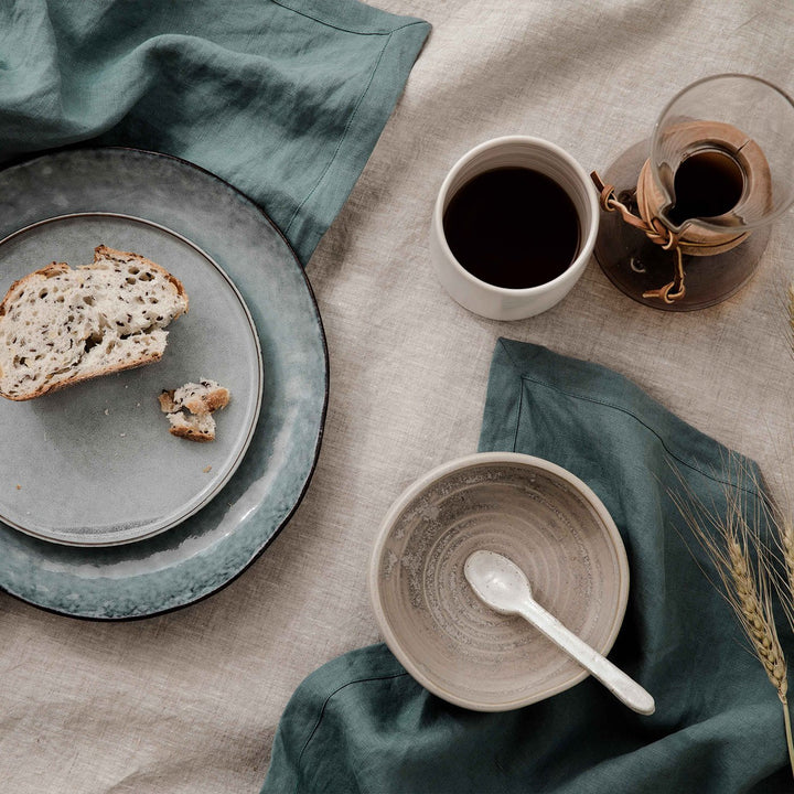 A table setting of a Linen Tablecloth in Natural, Linen Table Napkins in Bluestone and some ceramic plates with a piece of bread on top. There is also a jug of coffee and a mug on top of the tablecloth. 