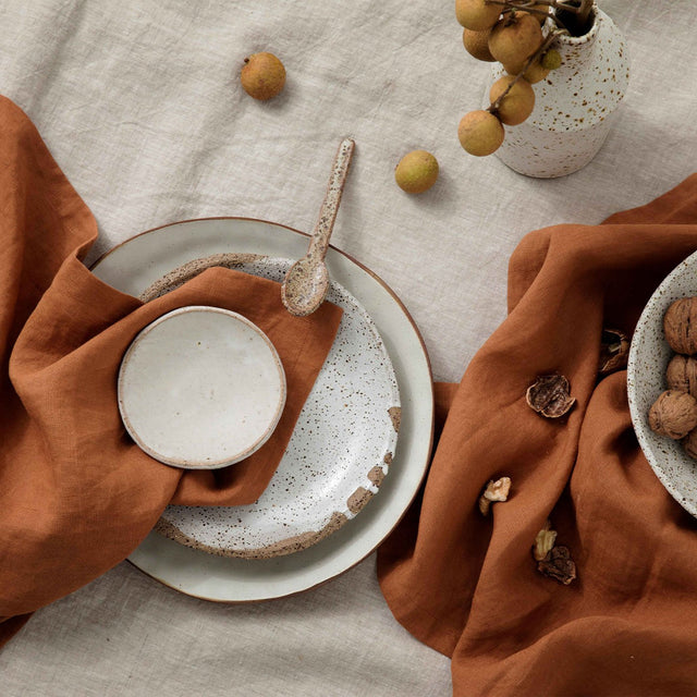 A table setting of a Linen Tablecloth in Natural and Linen Table Napkins in Cedar. There are various speckled ceramic bowls and plates on top of the tablecloth.