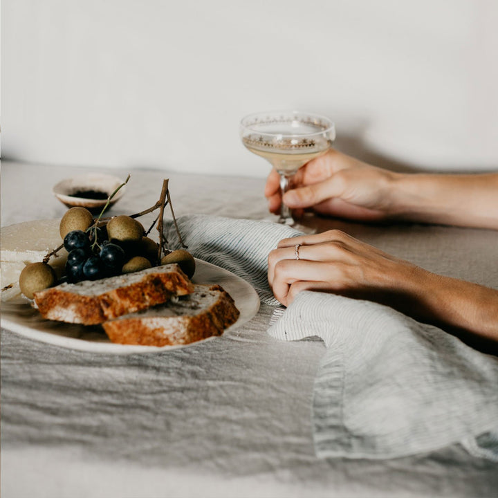 A table setting styled with a Linen Tablecloth in Natural, Linen Table Napkin in Pinstripe and a plate of bread and fruits. On top of the tablecloth are two arms reaching forward, one of which is holding a cocktail glass. 