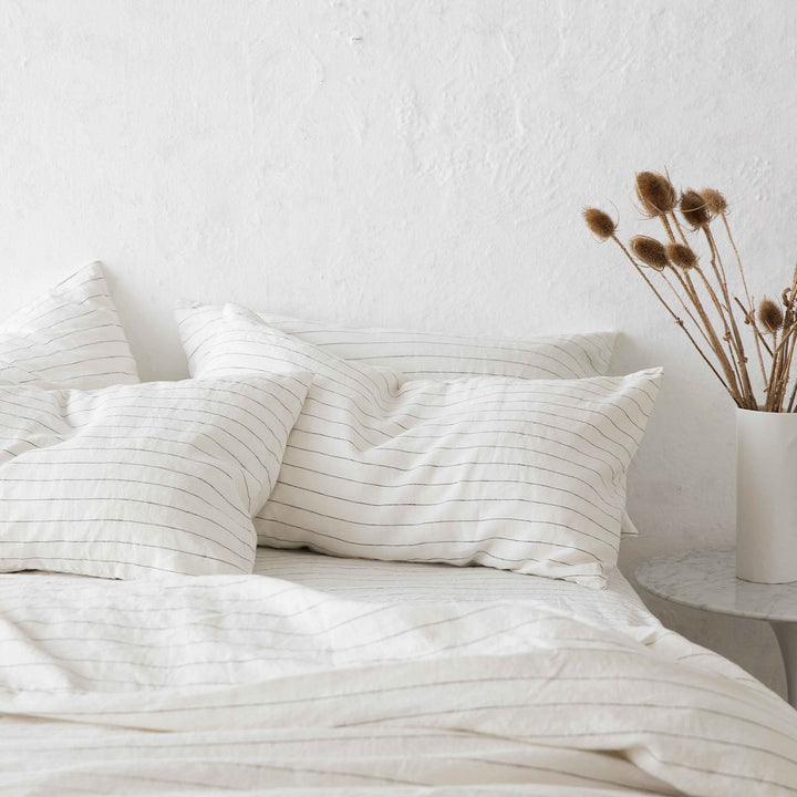 Set of 2 Linen Pillowcases and Linen Duvet Cover in Pencil Stripe on a bed.  Next to the bed is a marble bedside table with a white vase containing brown florals. Available in Standard & King.