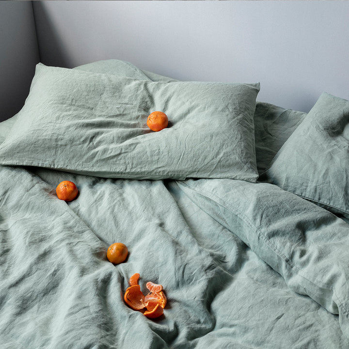 A bed dressed with linen bedding in Sage. On the bed are mandarins. Available in Double, King, Super King.