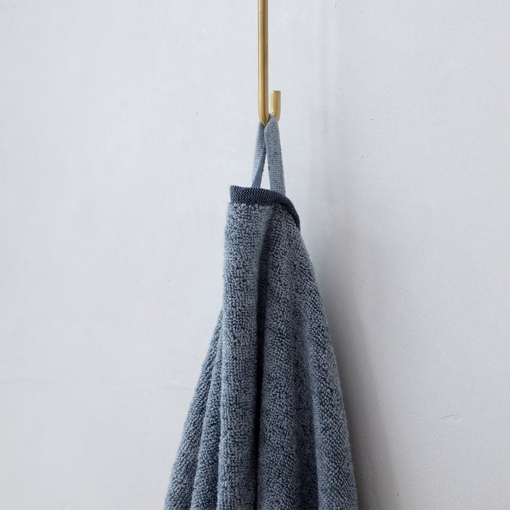  A close up photo of a Denim bath towel hanging on a hook. Available as Bath Towel Bundle & Bath Sheet Bundle. Available as Bath Towel Bundle & Bath Sheet Bundle.